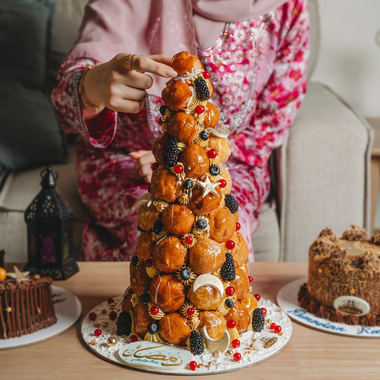 Person decorating a tall, ornate cake with fruit on a table.