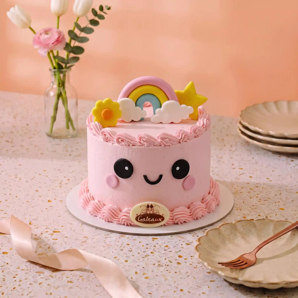 A pink frosted cake with a cute kawaii face, decorated with a rainbow, clouds, stars, and a yellow flower on top. The cake sits on a white plate on a speckled table with a peach-colored background, a vase of flowers, and plates.