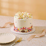 A frosted mini cake with white buttercream, colorful sprinkles, and piped cream on top, displayed on a speckled table with a plate, fork, and ribbon in a warm indoor setting.