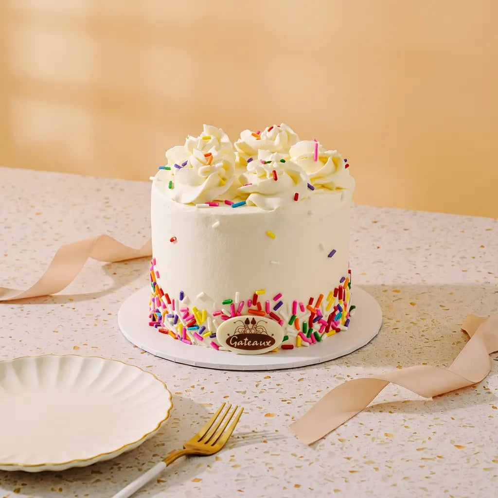 A frosted mini cake with white buttercream, colorful sprinkles, and piped cream on top, displayed on a speckled table with a plate, fork, and ribbon in a warm indoor setting.