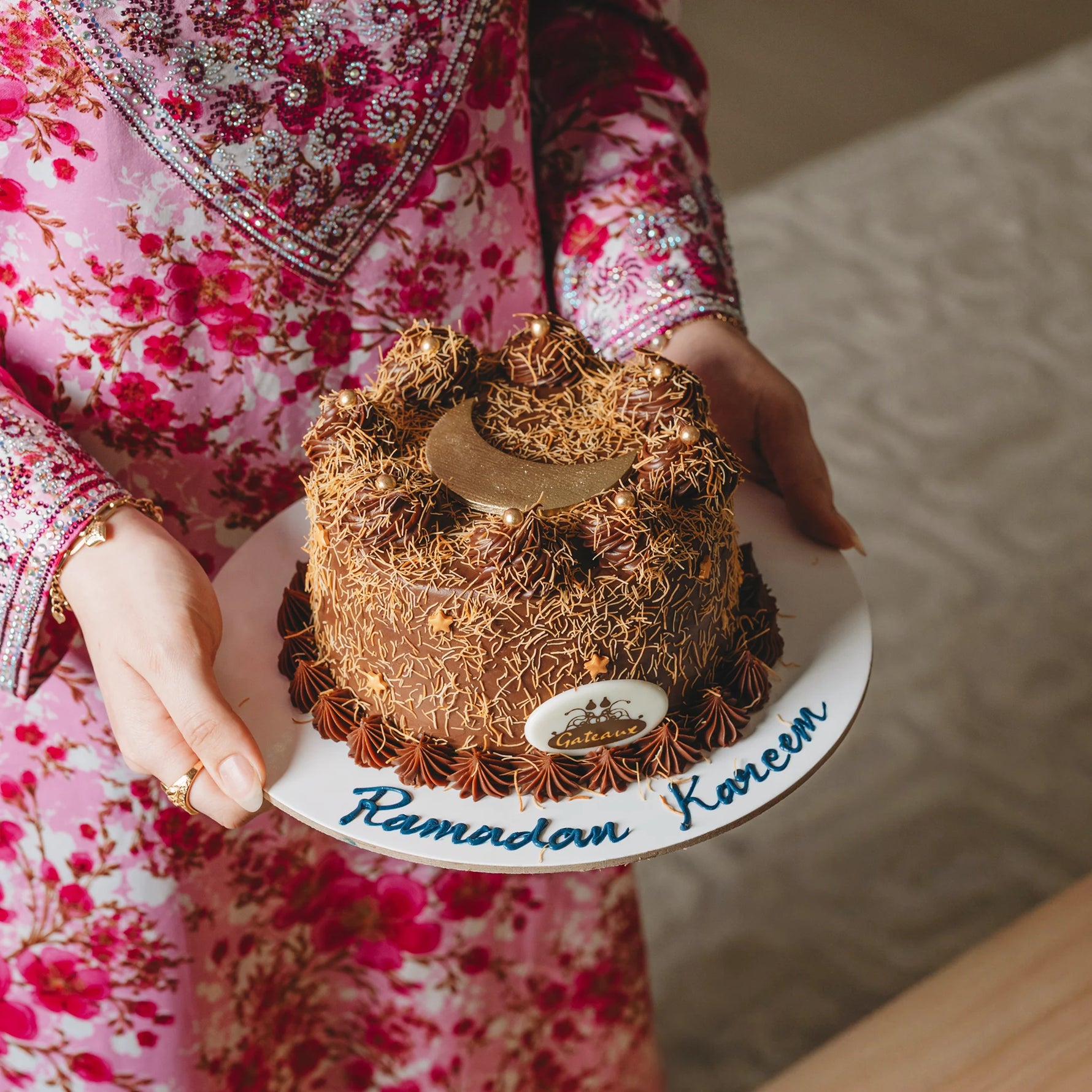 Person holding a decorated cake with 'Ramadan Kareem' text, wearing a pink floral outfit.