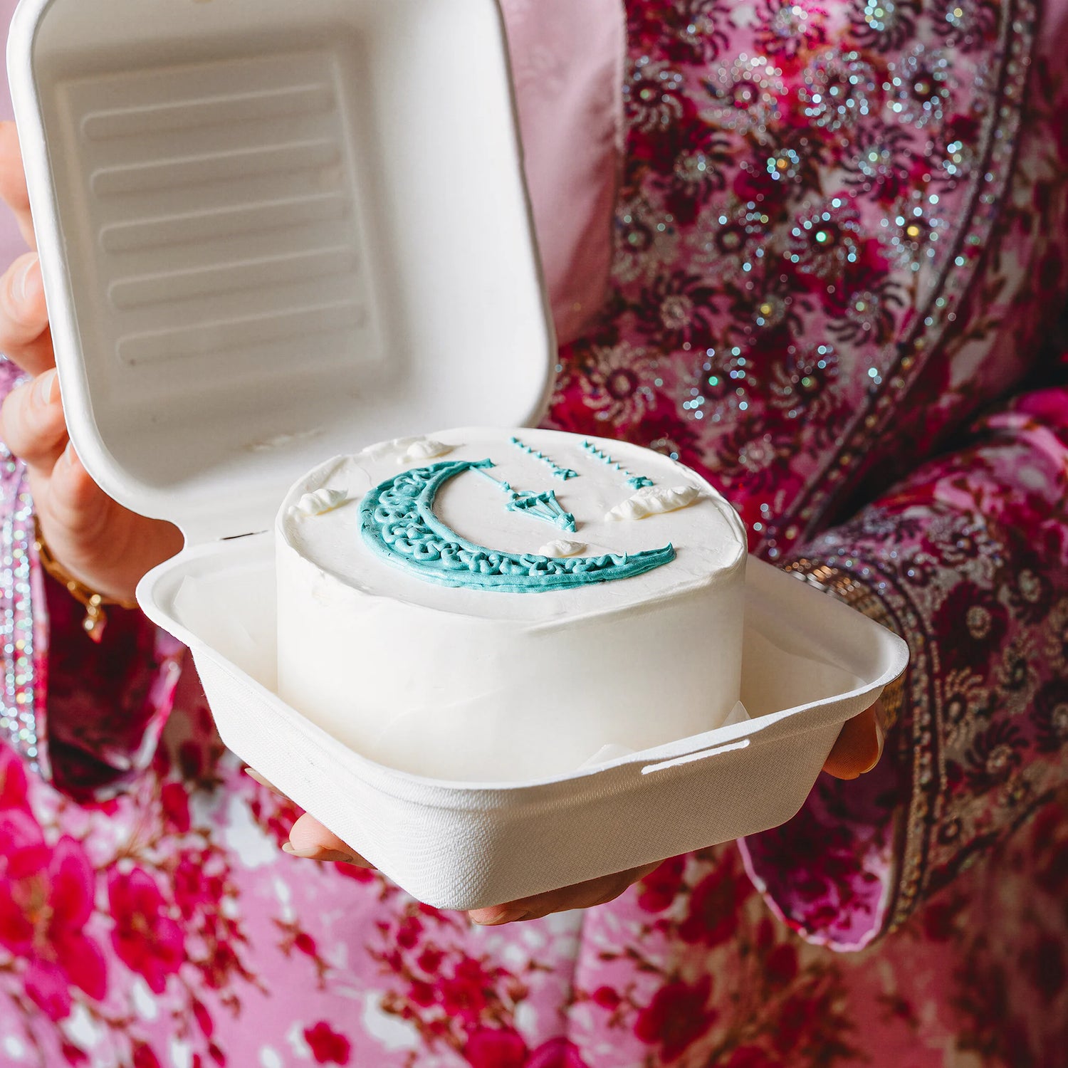 Person holding a small cake with blue icing in a takeout container, wearing a pink floral dress.