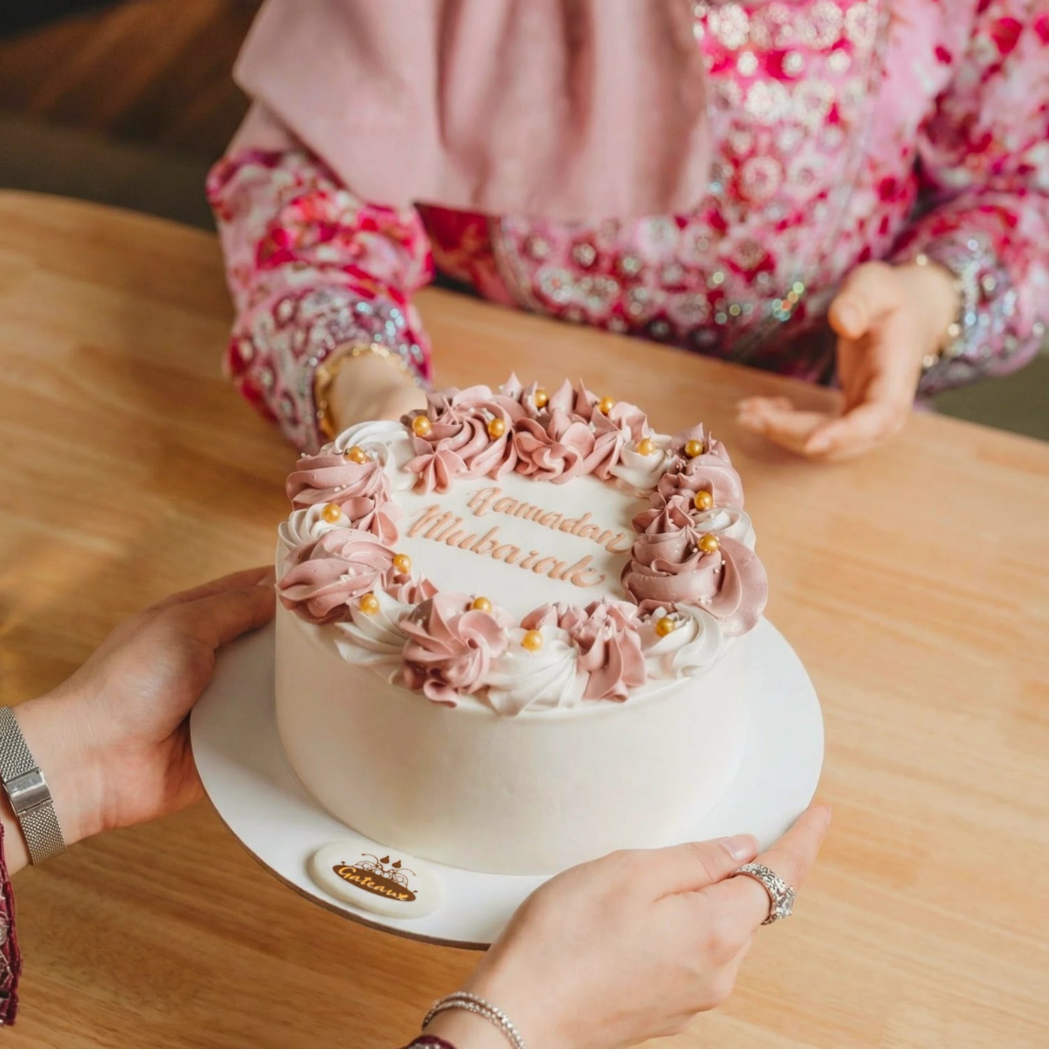 Decorative cake with pink frosting and gold accents on a wooden table, with a person in a floral dress partially visible.