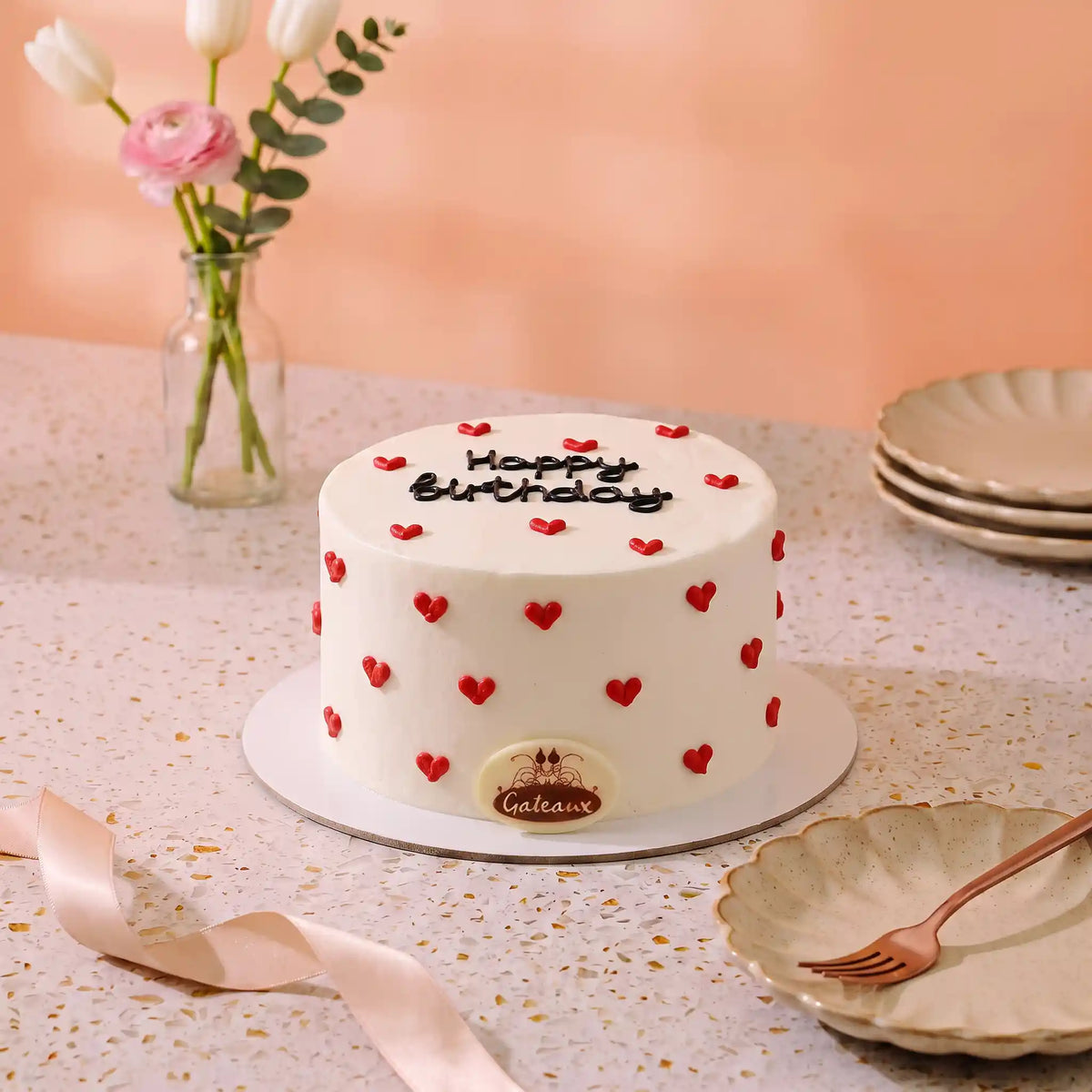 A white frosted birthday cake with red heart decorations and 'Happy Birthday' written on top, displayed on a speckled table with plates, a ribbon, and flowers in the background.