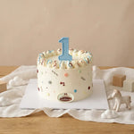 A round white birthday cake decorated with piped frosting, colorful confetti dots, and a large blue number 1 candle on top, displayed on a wooden table with a white cloth and small decorative props.