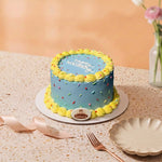 A round mini cake with light blue frosting, yellow piped border, and colorful sprinkles, displayed on a speckled beige table with a plate, fork, ribbon, and vase in the background.