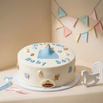 A round white cake decorated with blue and beige fondant details, including a blue baby outfit, small animal shapes, and the words 'Welcome to Baby Boy.' The cake sits on a white stand in a styled indoor setting with pastel bunting, a rocking horse, and ribbon accents.