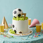 A white frosted birthday cake decorated with soccer balls, green grass-like frosting, a gold number 7, and gold 'Happy Birthday' script, styled on a table with party cups, cones, and streamers against a light blue background.