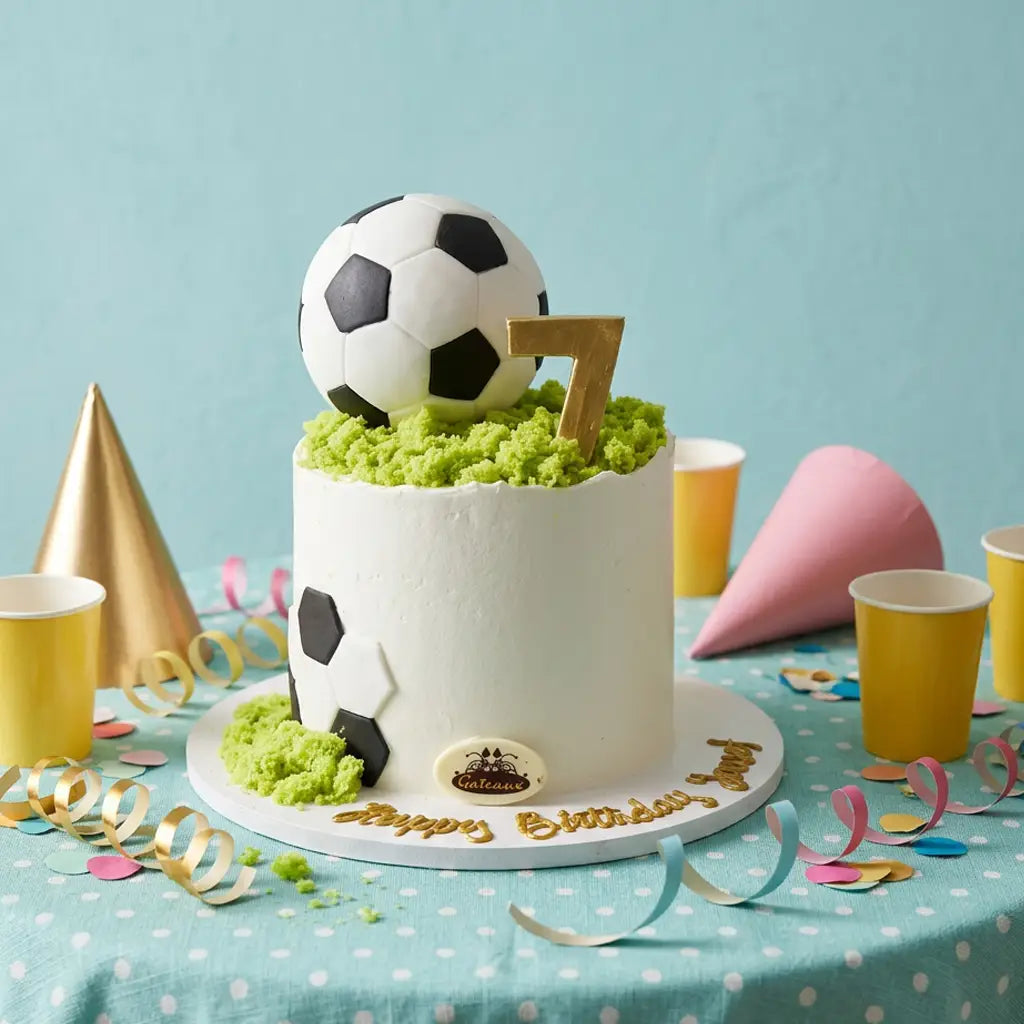 A white frosted birthday cake decorated with soccer balls, green grass-like frosting, a gold number 7, and gold 'Happy Birthday' script, styled on a table with party cups, cones, and streamers against a light blue background.