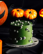 A Halloween-themed cake with a green layer and decorative spikes on top, placed on a table with pumpkins and a 'Happy Halloween' sign.