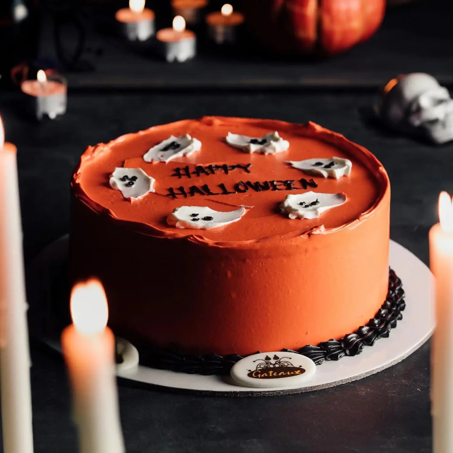 Halloween cake with orange frosting, black text, and white ghosts on a dark background with candles and pumpkins.