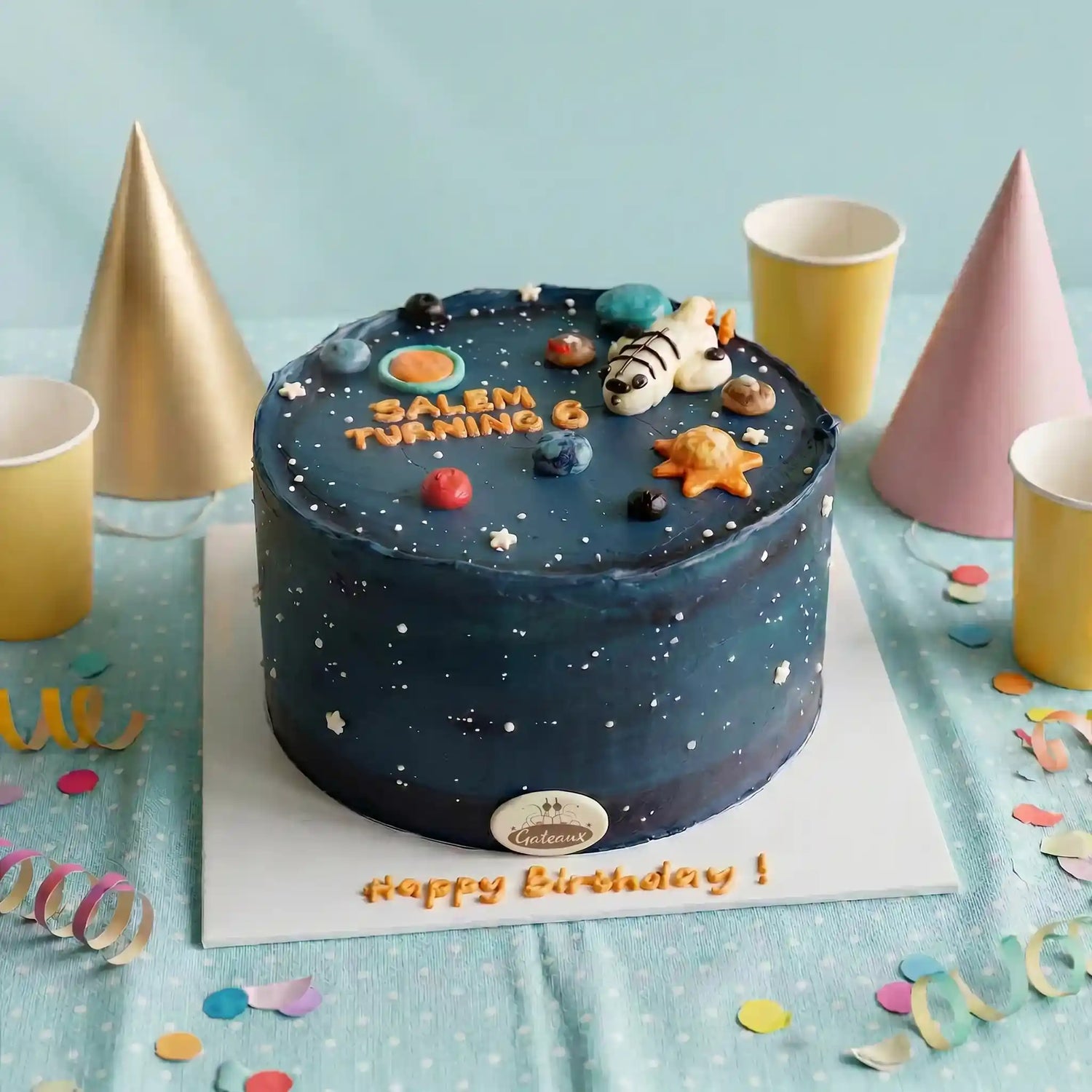 A round birthday cake decorated with a dark blue space theme, featuring small white stars, colorful planets, a seashell, and the text 'SALEM TURNING 6' and 'Happy Birthday!'. The cake sits on a white board surrounded by gold, pink, and white party cups and confetti on a light blue fabric backdrop.