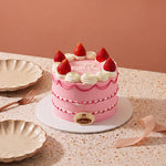 A pink frosted mini cake decorated with fresh strawberries and white whipped cream, displayed on a speckled beige table with decorative plates and a ribbon.