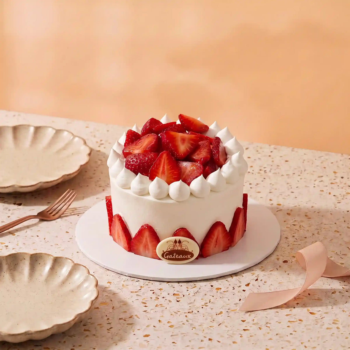 A strawberry cake on a white round cake stand, decorated with fresh sliced strawberries on top and around the sides, set on a speckled beige table with plates and a fork in the background.