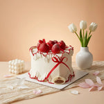 A white frosted square cake decorated with fresh strawberries, red ribbon bows, and a small gold Cartouche plaque, styled on a beige surface with tulips in a vase and pink petals around it.