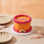 An orange round mini cake with pink piped frosting borders, pink heart decorations, and 'Happy Birthday' written on top, displayed on a white plate on a speckled beige table with plates and a ribbon nearby.