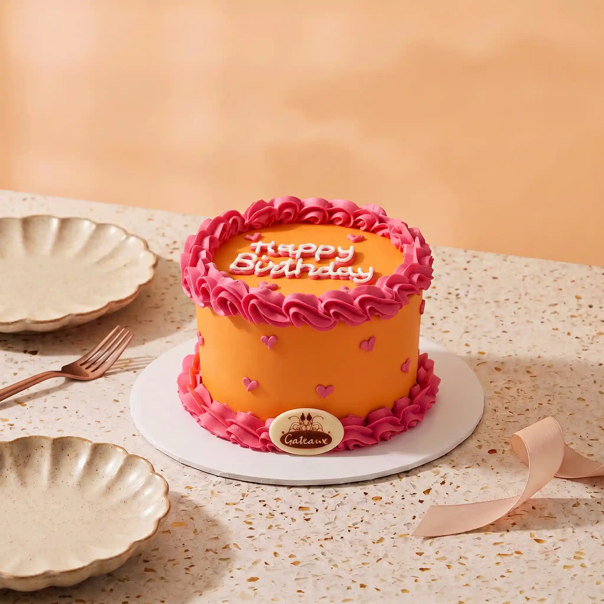 An orange round mini cake with pink piped frosting borders, pink heart decorations, and 'Happy Birthday' written on top, displayed on a white plate on a speckled beige table with plates and a ribbon nearby.