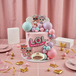 A pink LOL Surprise cake decorated with pastel balloons, character toppers, and a 'Cakes' sign, styled on a pink tablecloth with plates, gold foil accents, and gift boxes.