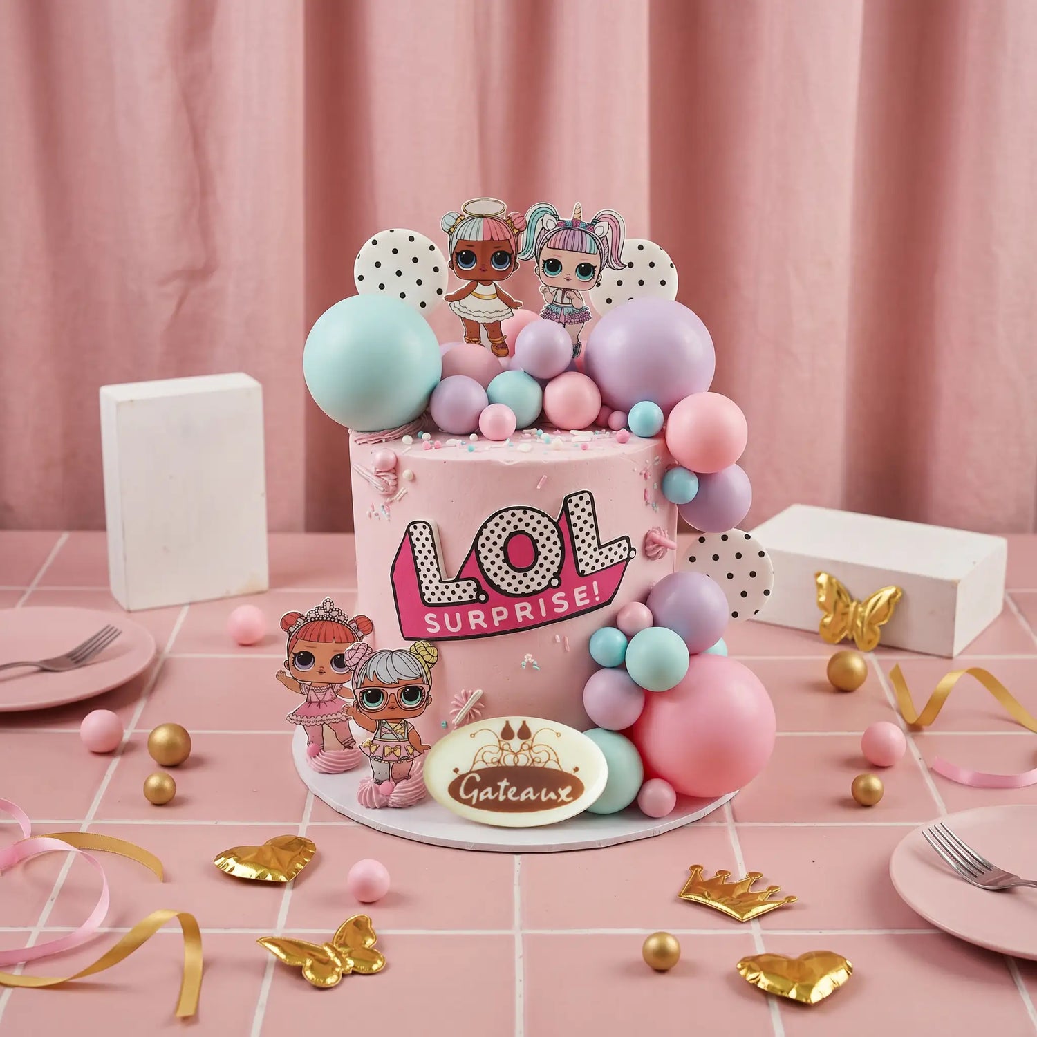 A pink LOL Surprise cake decorated with pastel balloons, character toppers, and a 'Cakes' sign, styled on a pink tablecloth with plates, gold foil accents, and gift boxes.