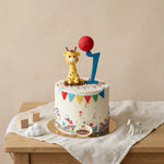 A decorated white round cake on a wooden table, topped with a yellow giraffe figurine holding a red balloon and a blue number 1 topper. The cake has colorful confetti, bunting, and a small round cake label reading 'Bakery'.