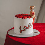 A Valentine's Day cake with white frosting and the word 'love' written on it, decorated with a red heart pattern and topped with a small teddy bear holding a heart.
