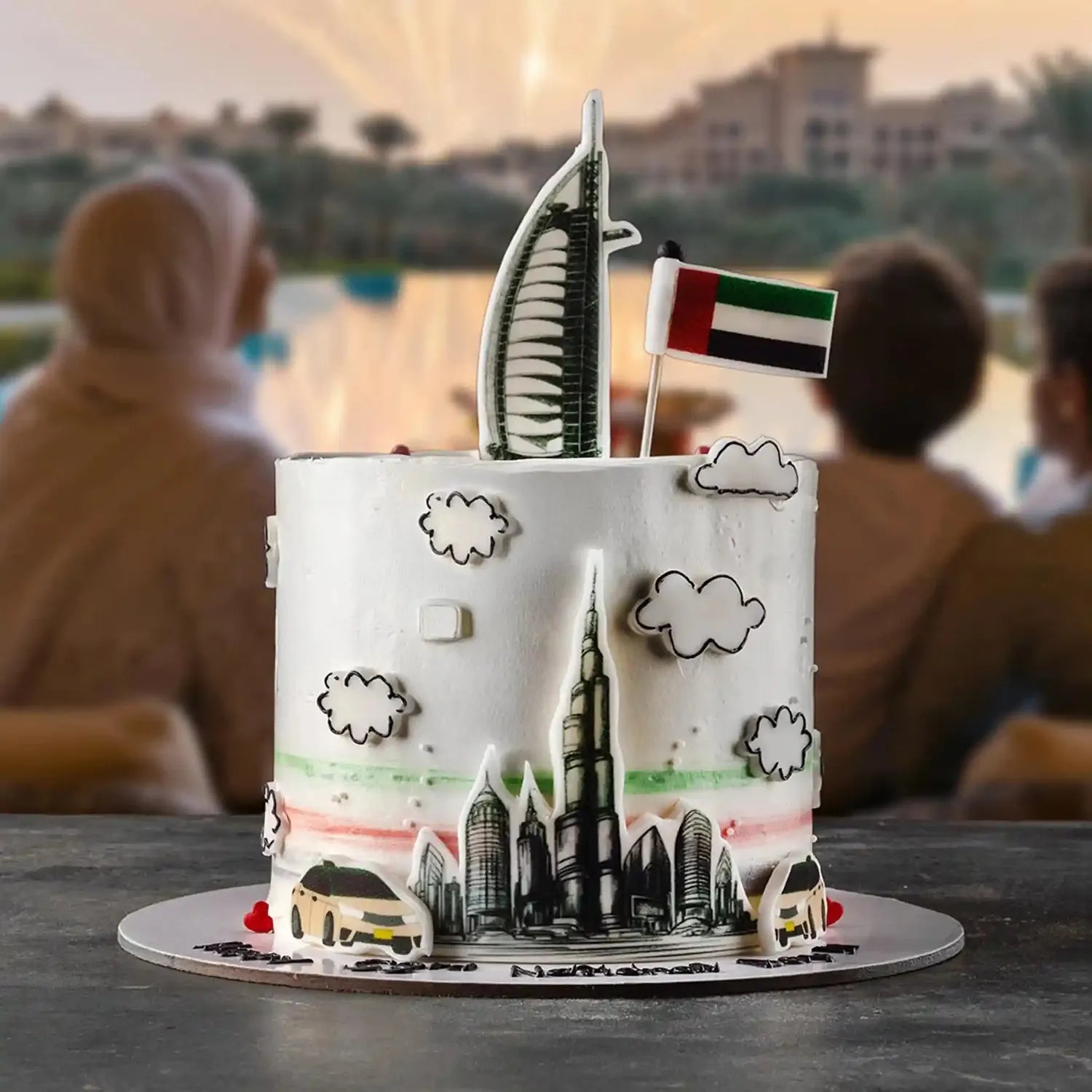 Cake with architectural designs and a flag, set against a blurred background of people and a scenic view.