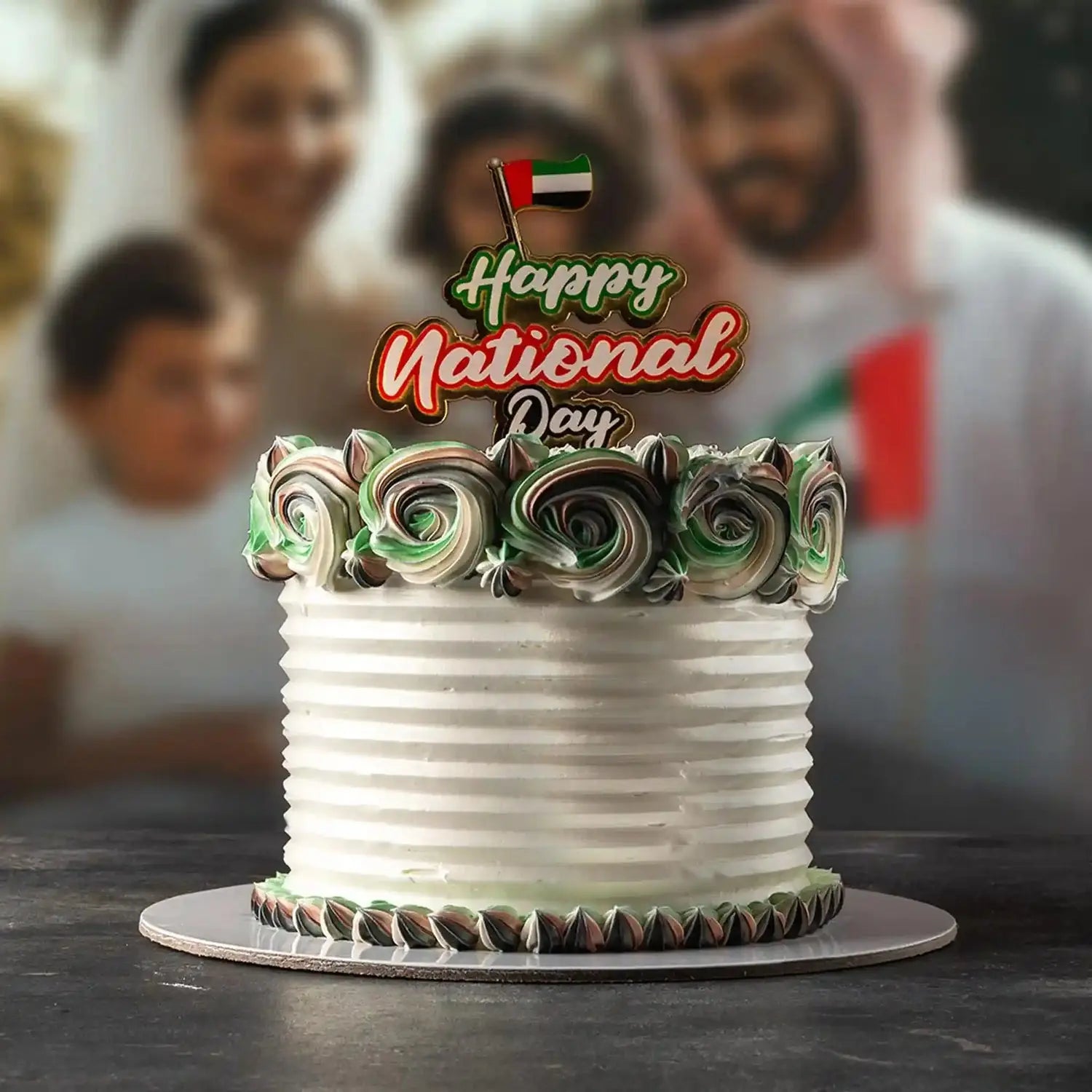 White cake with decorative swirls and 'Happy National Day' topper in front of a blurred background with people.
