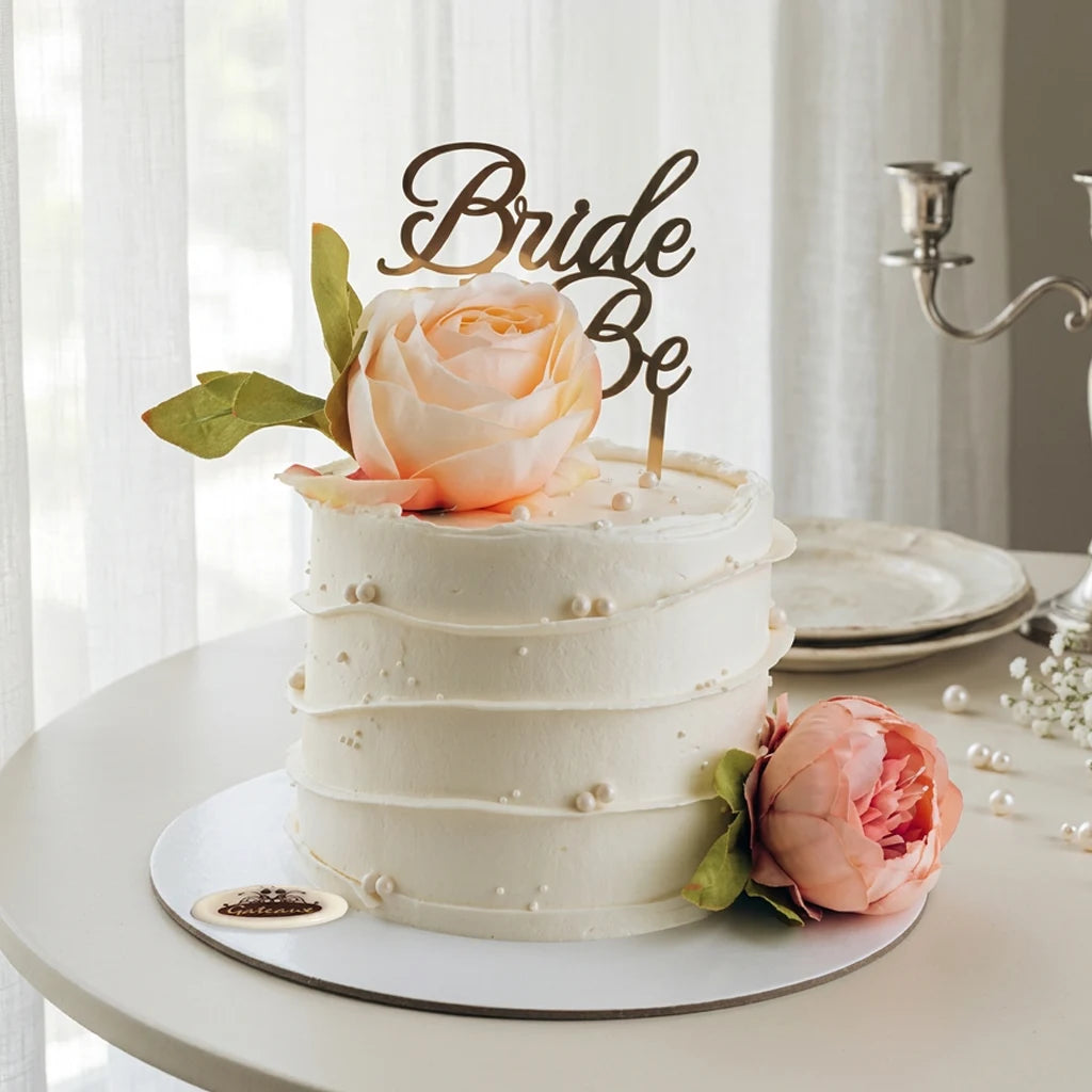 A frosted white tiered cake on a round table with a peach rose topper and a gold 'Bride & Be' sign, set in a softly lit indoor setting with plates and a candle holder in the background.