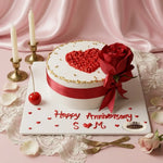 A round white cake decorated with a red heart, red ribbon, and a large red rose, placed on a white board with 'Happy Anniversary S M' written in red icing. The scene is styled with pink satin fabric, candles, lace, petals, and forks.