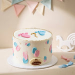 A round white frosted cake decorated with pink and blue footprints, colorful heart shapes, and a gold question mark on top, displayed on a white cake board against a warm cream background with pastel bunting and a white rocking horse decoration.