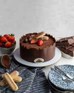 A chocolate Tiramisu Cake decorated with strawberries on top, with ladyfinger biscuits and a bowl of strawberries in the background.