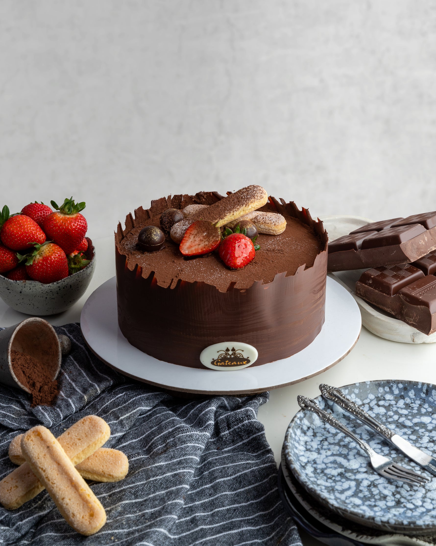 A chocolate Tiramisu Cake decorated with strawberries on top, with ladyfinger biscuits and a bowl of strawberries in the background.