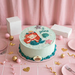 A round birthday cake decorated with a mermaid design, pink starfish, teal seashells, and the words '3 years' on a pink tablecloth, surrounded by white boxes, plates, and gold and pink decorative props.