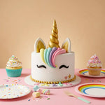 A white unicorn-themed cake with a gold horn, pastel rainbow mane, and white ears, styled on a pink table with cupcakes, rainbow plates, and sprinkles against a peach background.