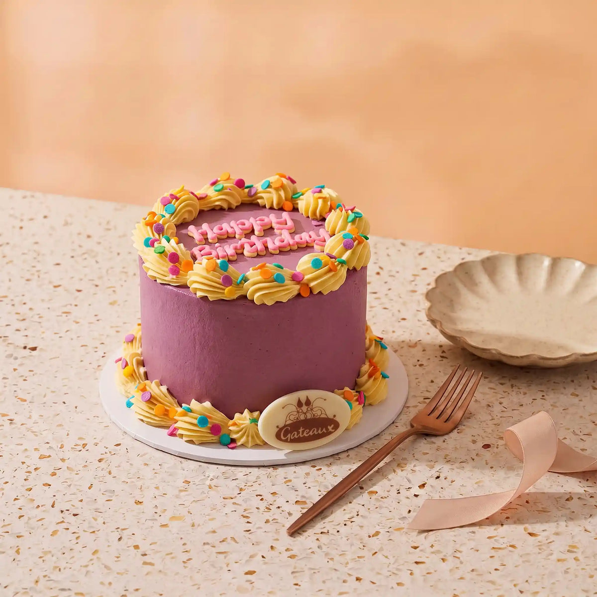 A round mini cake with purple frosting, yellow piped borders, colorful sprinkles, and pink 'Happy Birthday' lettering on top, displayed on a speckled tabletop with a small plate, fork, and ribbon nearby.