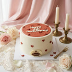 A round birthday cake decorated with white frosting and a reddish-brown top layer, topped with the words 'Happy 4th Anniversary' and small red hearts. The cake is placed on a white table runner with pink roses, candles in brass holders, and lace around it.