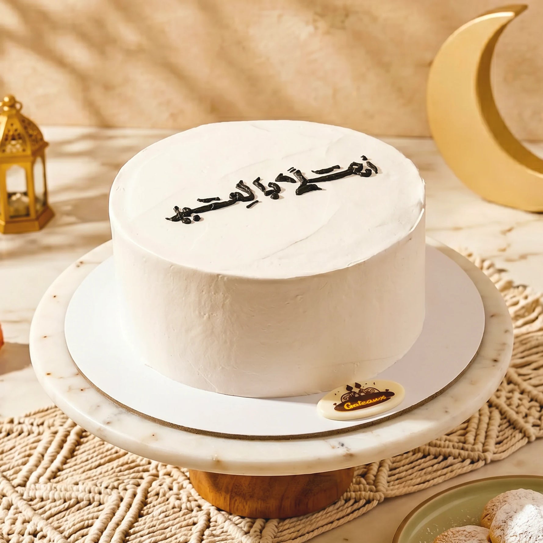 White cake with Arabic text on a decorative stand with a gold crescent moon and lantern in the background.