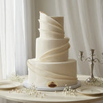 A three-tier elegant wedding cake with smooth white fondant and decorative spiral ridges, displayed on a round table with plates, pearls, and a silver candelabra against a soft white curtain background.