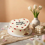 Heart-shaped white frosted cake with red heart decorations and 'happy one year' written on top, displayed on a marble surface with tulips in a vase and a candle nearby.