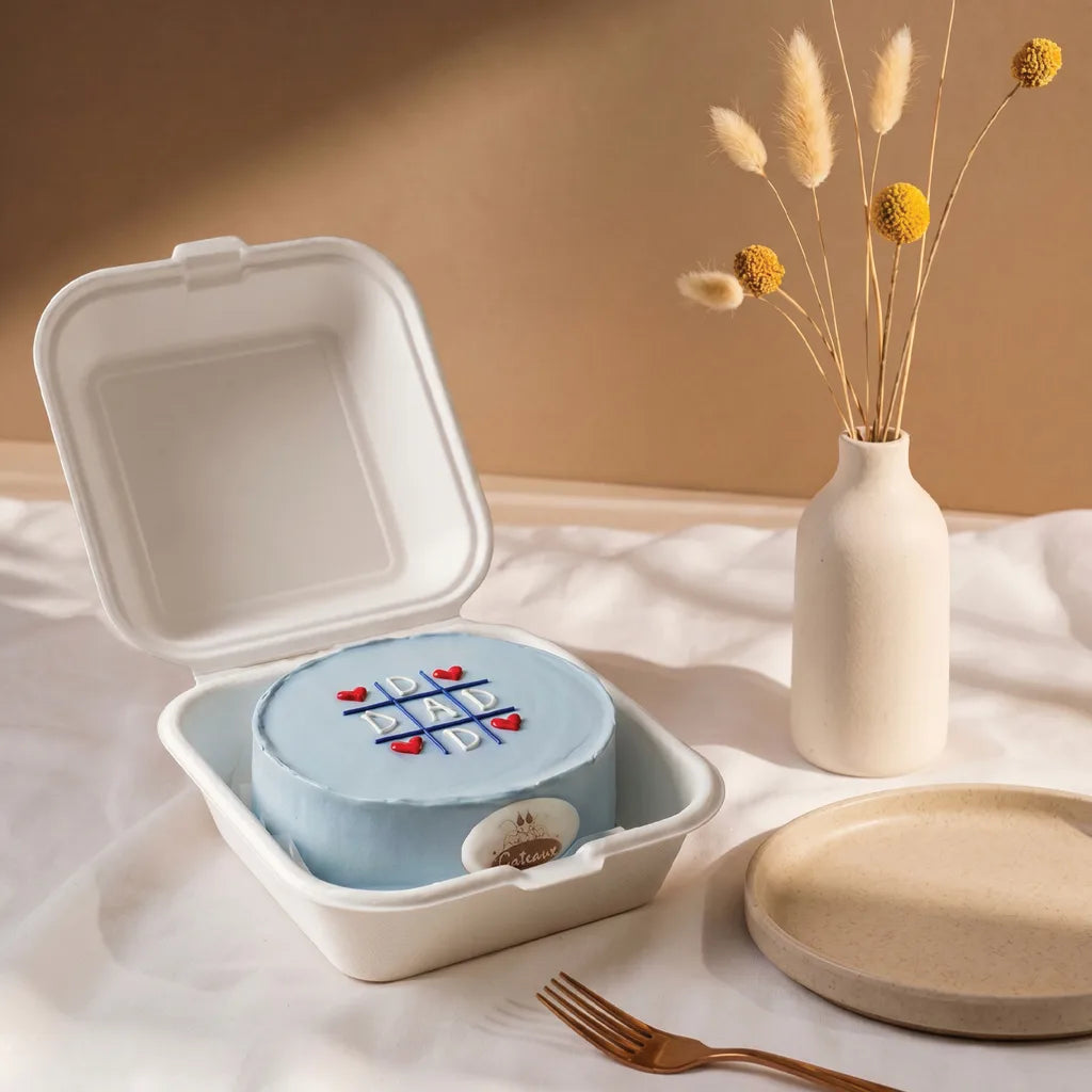 Small blue cake in a takeout container on a white surface with a vase of dried flowers in the background.