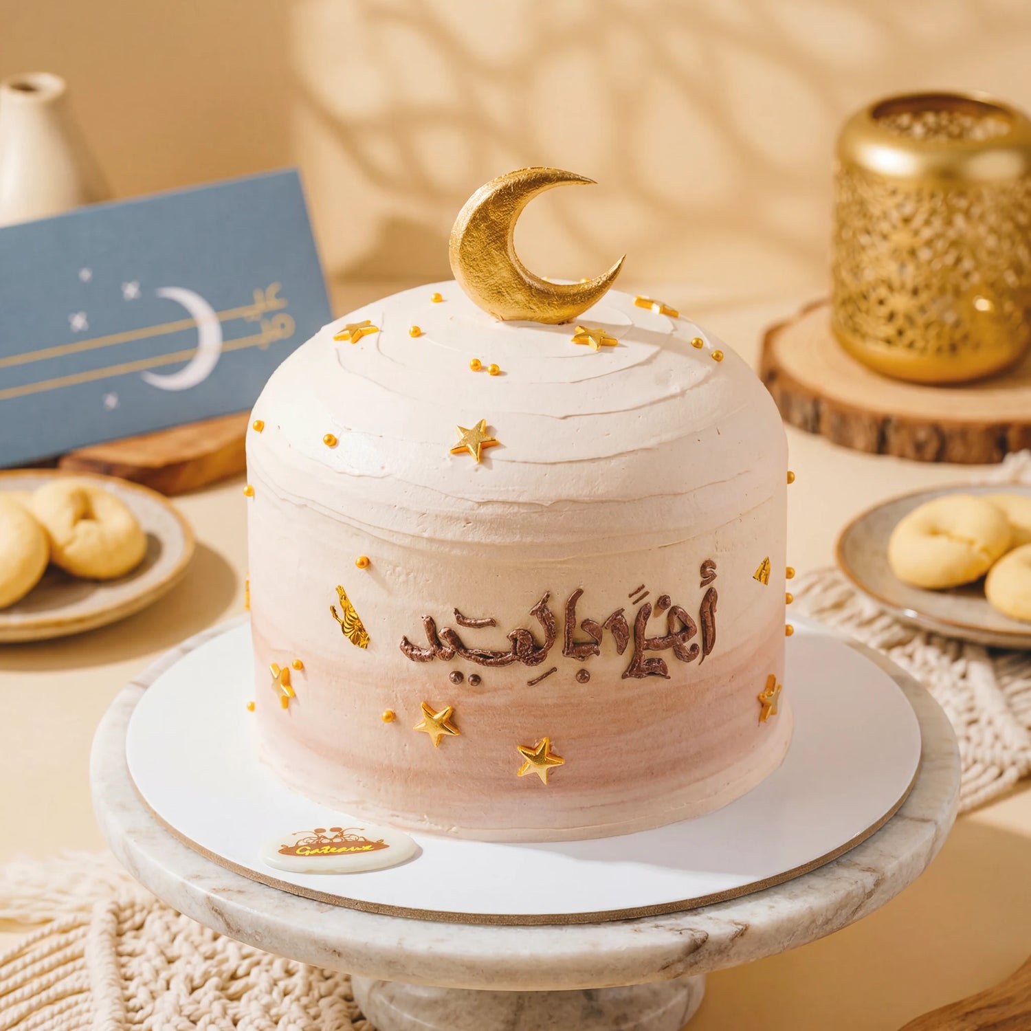Decorative cake with gold accents on a marble stand, surrounded by cookies and a card.