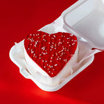 Heart-shaped red cake with white frosting in a white box on a red background