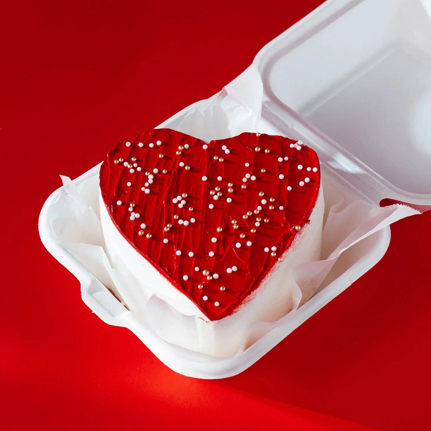 Heart-shaped red cake with white frosting in a white box on a red background