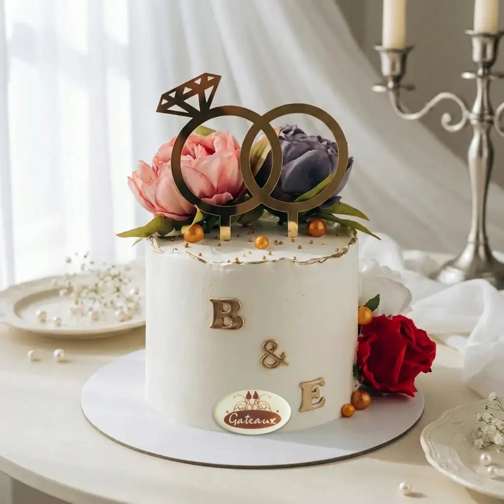 A white frosted birthday cake decorated with gold ring-shaped toppers, pink and purple peonies, a red rose, and gold lettering spelling 'B & E', shown on a round cake board in a softly lit indoor setting.